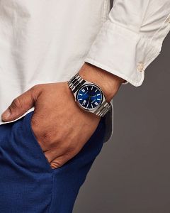 Hand wearing a silver watch with a blue face against a gray background